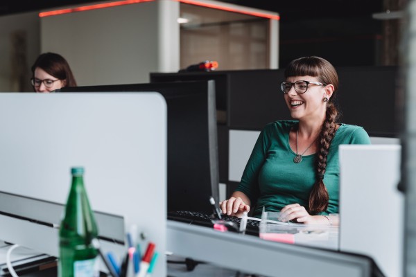 Zwei Personen sitzen nebeneinander an ihren Arbeitsplätzen vor Computerbildschirmen. Die Person im Vordergrund lacht.