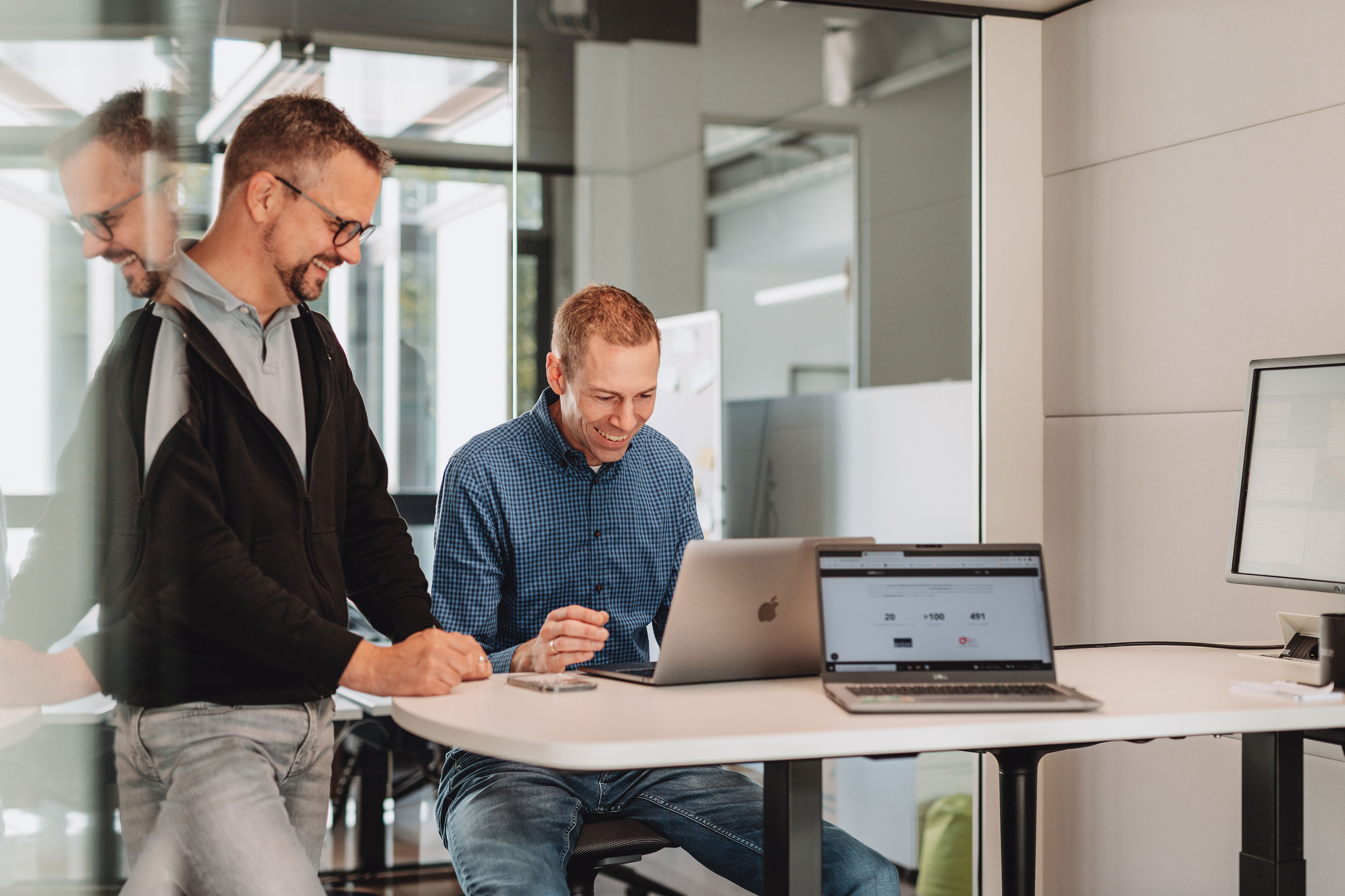 Eine Person sitzt an einem kleinen Konferenztisch an einem Notebook. Eine Person steht daneben, beide schauen gemeinsam auf den Monitor den Notebooks.