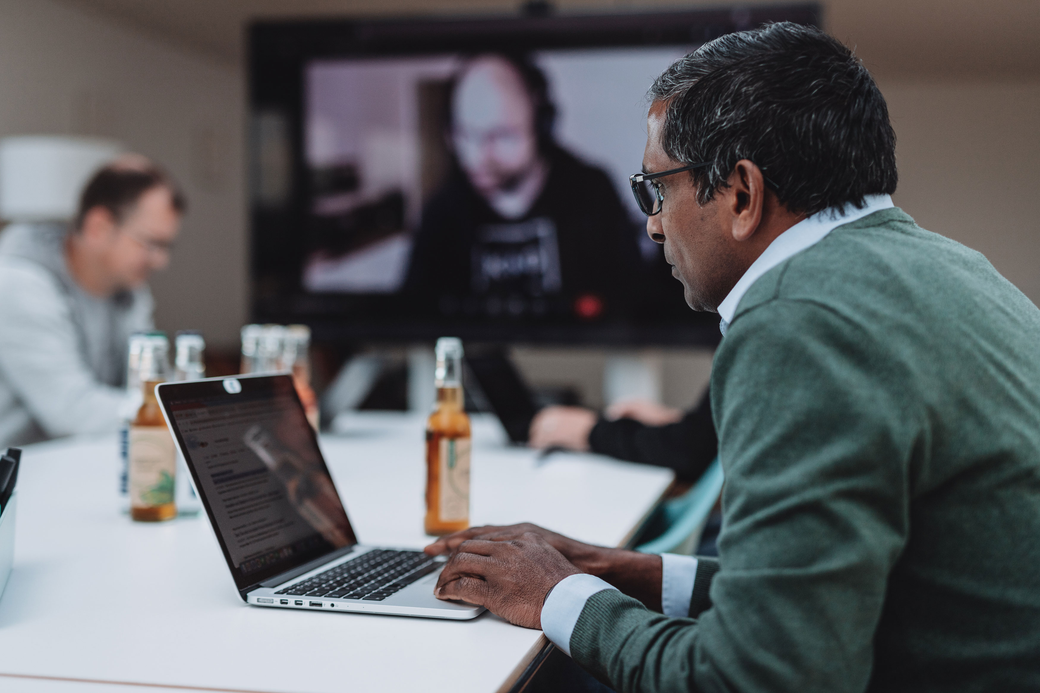 Eine Person arbeitet an einem Notebook in einem Konferenzraum. Im Hintergrund steht ein Bildschirm, auf dem eine weitere Person in einer Videkonferenz zu sehen ist.