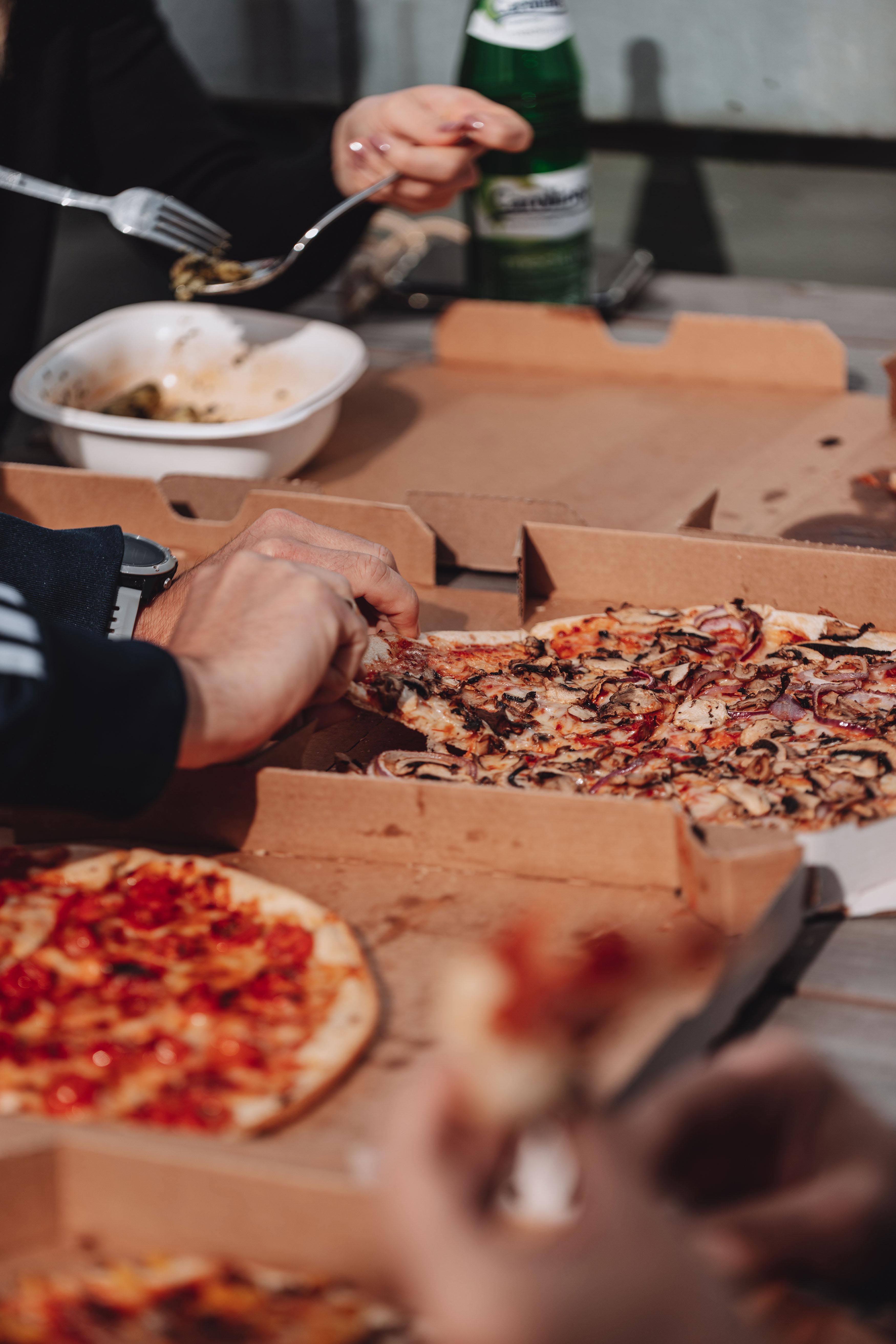 Pizzen liegen in ihren aufgeklappten Kartons auf ihrem Tisch. Eine Hand greift nach einem Stück Pizza.